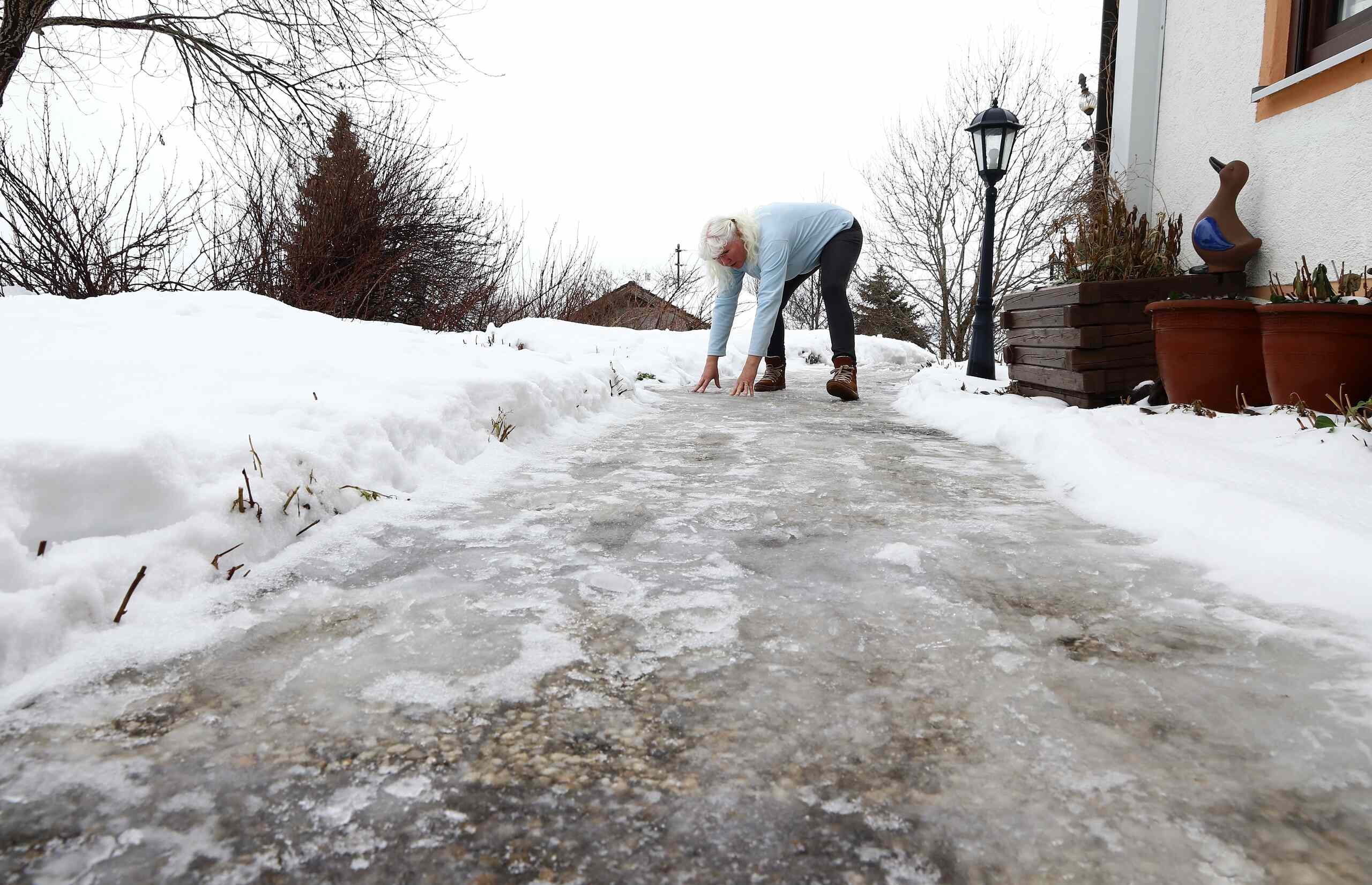 A close-up side view of a person's legs in black boots walking cautiously across a dark brick pavement covered in patches of melting ice and slush, representing pedestrian hazards evaluated in Springfield winter slip & fall cases.
